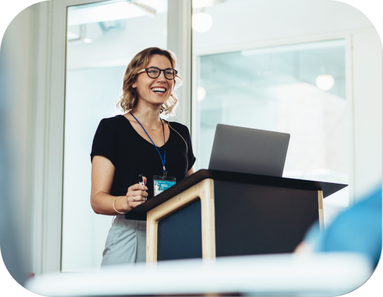 mi1 Confident female speaker giving a presentation at a conference, standing at a podium with a laptop.
